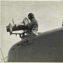 A close-up view from low left side of nose of aircraft, looking forward, of a man in a flight cap that stands in the forward cockpit of a Naval Aircraft Factory F-5L (PN-5) demonstrating a hand-operated K-2 aerial camera used for oblique aerial photography.