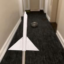 A paper rocket on a straw in the foreground. A colander on carpet in the background.