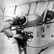 A man reaches out from the back seat of a biplane to look into the viewfinder of a camera that is strapped to the side of the aircraft.