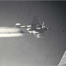 Grainy image of an aircraft flying with a trail of contrails.