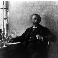 A portrait of a man sitting in a chair at a desk, facing the viewer. 