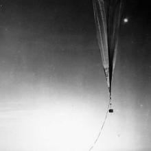 A balloon floats just above the Earth's surface with a camera attached to it. A group of people stand in a semi circle on the ground below it.