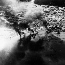 An aerial photograph of a small city with large plumes of smoke coming up.