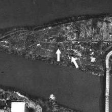 An aerial photo of a triangular shaped island that has a bridge connecting it to the mainland. Arrows points to dark patches near the center of the island.