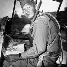 Photograph of a man sitting in an aircraft holding a map and looking toward the camera.