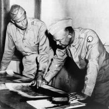 Two men in military uniforms stand at a table looking at photographs.