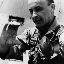 A man in a flight suit turned three-quarters of the way toward the camera, he makes a c-shape with his hand.