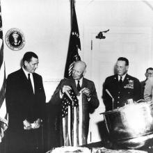 A group of 5 men stand around a table with a large metallic drum shaped object on it.