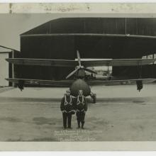 Black and white panoramic photograph of five airplanes, each plane has two men in flightsuits standing in front, with a hangar in the background. There are folds throughout the image.