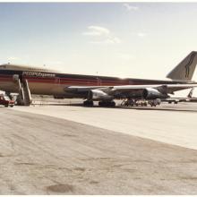 Color photograph of jet aircraft with red white and blue stripes and the text "PeoplExpress" on the fuselage. A stylized drawing of two parallel faces in profile are on the tail.