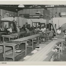 Black and white photograph of corrugated metal pieces with men and women working in the background