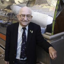 Balding elderly man wearing glasses stands beside a silver reflective airplane nose.