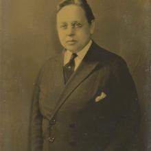 Sepia-toned photo of a woman wearing white collar, dark Windsor tie held with a stick pin, and a double breasted coat