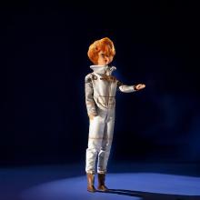 The Miss Astronaut doll photographed against a dark background. She wears a silver spacesuit and has a short haircut.