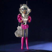 1985 Astronaut Barbie photographed against a dark background. She wears pink boots, silver leggings, a poofy pink top, and a space helmet.