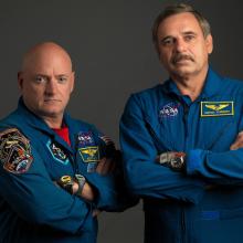 Two astronauts in blue jump suits with their arms crossed stare into camera. 