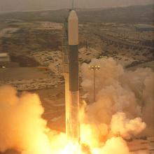 A rocket launching off from the ground. Plumes of smoke and fire combust, pushing it upward.