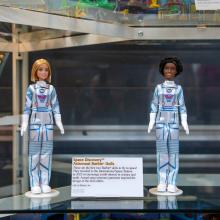 Two Space Discovery Barbies photographed in a glass case. They appear to be standing up straight. Between them is a small artifact description card.