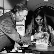 A black and white photograph of a young woman showing a maze to an older man in a suit.