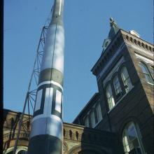 The top three-quarters of green and white missile seen from the ground looking up. The rocket sits vertically in front of an ornate building.