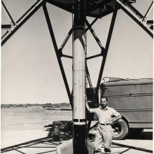 A man leans on a rocket that is about two times as tall as him. The rocket is fixed to a launch tower. Part of a truck can be seen in the background.