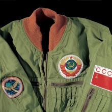 The top half of a green synthetic, woven cloth coverall with burnt orange collar.  The suit has a Soviet flag patch on the left shoulder with the letters CCCP along the top; a patch of the Soviet seal above the left breast pocket; and on the right shoulder is a Salyut space station program patch.