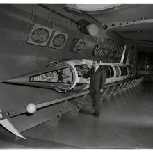 A man stands with one hand on a rocket laying horizontally in a hallway.