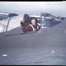 A woman in a military uniform with 1940s styled hair waves from the cockpit of an airplane?