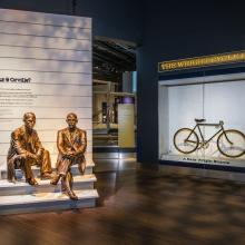 A photo looking into the entrance of a museum exhibit. A bicycle can be seen on display behind plexiglass and a statue of two men sitting adorn the entryway panel which says "who were wilbur & orville."