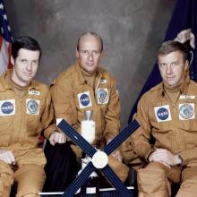 Three people in orange jumpsuits sit posed in front of two flags with a small model of Skylab in front of them.