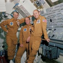 Three men stand facing the camera. The two on the righthand side lean against a control board inside a training module at NASA.