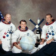 Three people seated against a neutral background with an American flag to the left. All three people are wearing spacesuits. The person in the middle holds a globe. A model of Skylab is situation between the person in the middle and the person on the right.