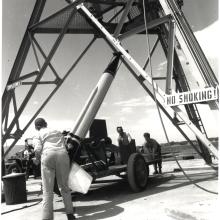 A group of men push a wheeled vehicle underneath a large tower. A rocket sits atop the wheeled vehicle. A sign that says "no smoking!" is attached to the tower.