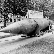 A large rocket lays horizontally on a city street with ropes around it. A sign on top of it states that it is a captured German V-2 rocket.