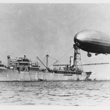 An image of a military ship at sea with a mooring mast. An airship is docked at the mast.