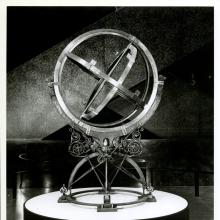 A black and white photo of a spherical sculpture with three sets of metal rotating rings. The center ring is the smallest and each ring after encompasses the one before it.