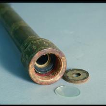 A photo of the end of a simple wooden telescope embellished with golden filigree painted on its surface. The lens and end cap are laid on the table next to it.