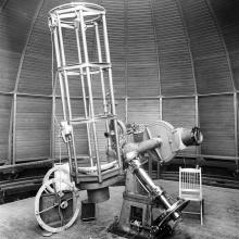 A black and white photograph of a large telescope inside of an observatory. A chair is situated next to it, the juxtaposition demonstrating that the chair is roughly 1/3 the size of the chair.