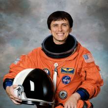 An astronaut holding a helmet in his hand and wearing a bright colored spacesuit smiles for a portrait.