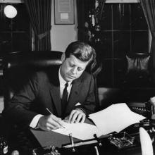 President Kennedy leans over a desk signing a document. 