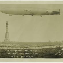 An airship floats in the air at the top of the frame. A mooring mast can be seen in the distance behind it.