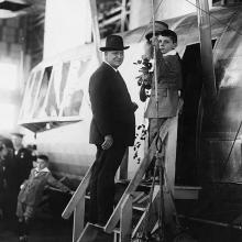 Three people stand on steps that lead to the entrance of an airship control car. They all look to the left, facing the camera.