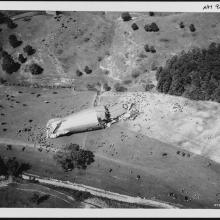 A cylindrical piece of an airship broken up in a field as seen from above. People crowed around it.