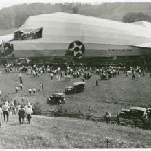 A wrecked piece of an airship sits in a fields as swarms of people flock to it.