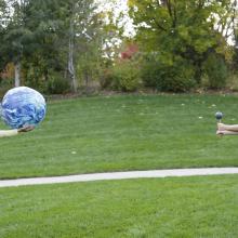 Two children stand on the grass about 6 feet away from each other. One holds a ball that represents the Earth. One holds a ball that represents the Moon. 