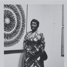 An African American woman stands proudly in a 1970s dress in front of an abstract painting.