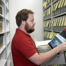 A man opens an archival box and holds a news magazine. On the magazine is a picture of Earth from space. Behind him are shelves of archival boxes. 