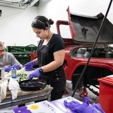 Two interns, a young man and young woman, work at a table wearing gloves and eye protection. Behind them is a jeep with the hood open. 