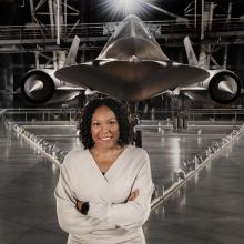 A woman stands in front of a large black airplane.