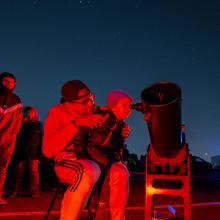 An child looks through a telescope with a thrilled expression while sitting on an adult's lap.